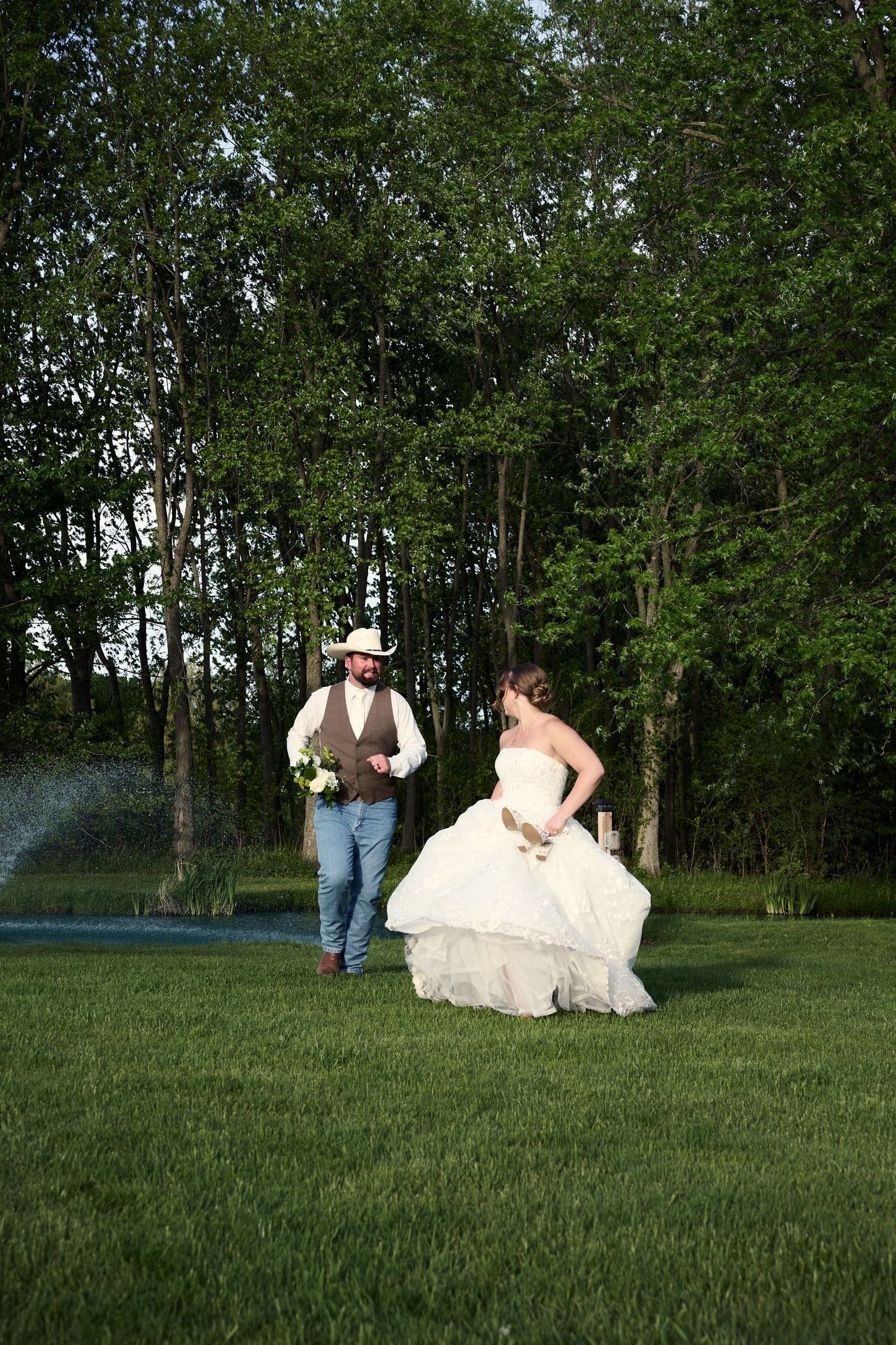 Bride in white gown and groom in cowboy hat walking together on green lawn at Crescent Hill Acres wedding in Canada
