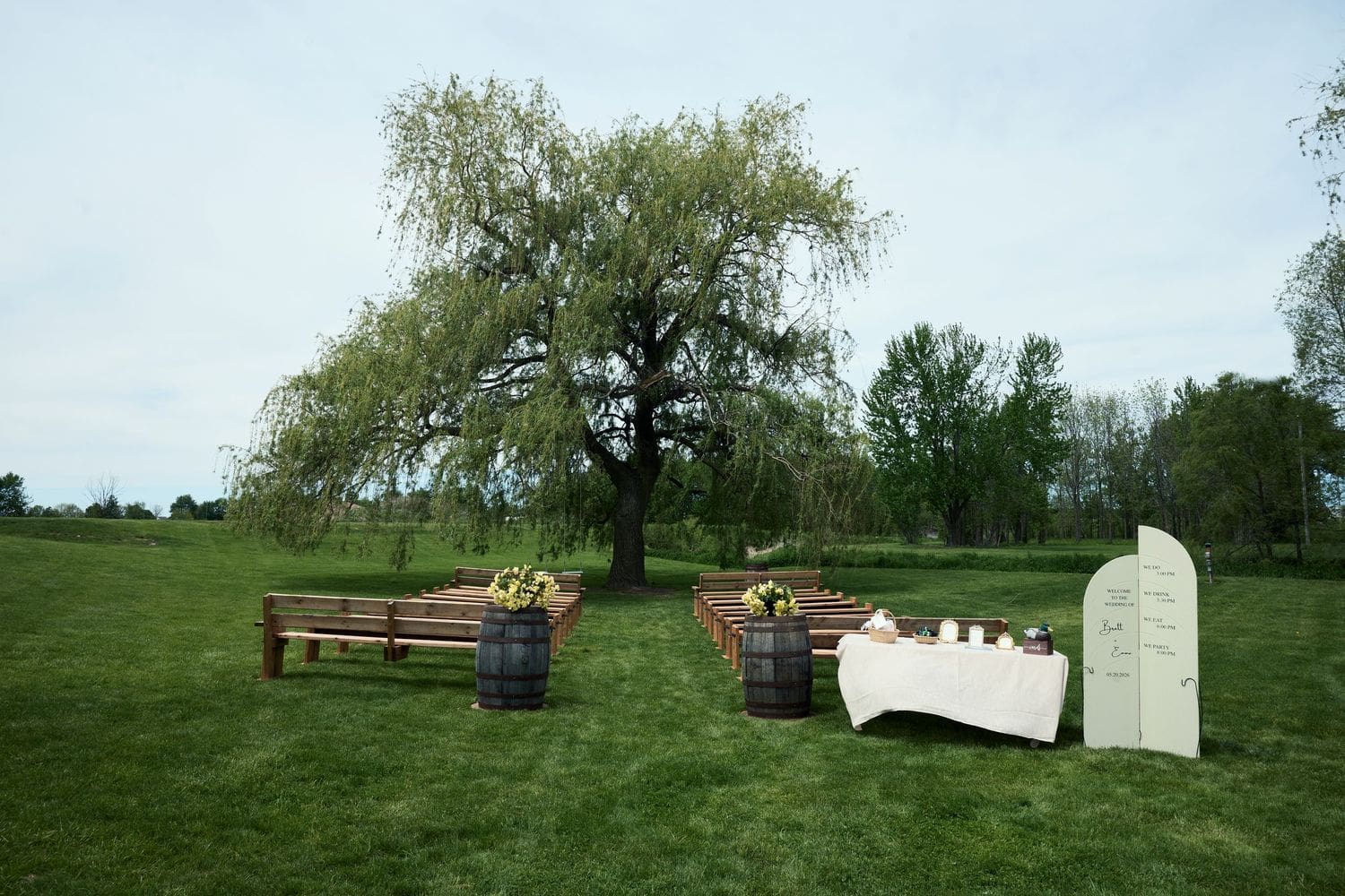 Rustic outdoor wedding ceremony setup with wooden barrels and benches beneath a weeping willow at Crescent Hill Acres