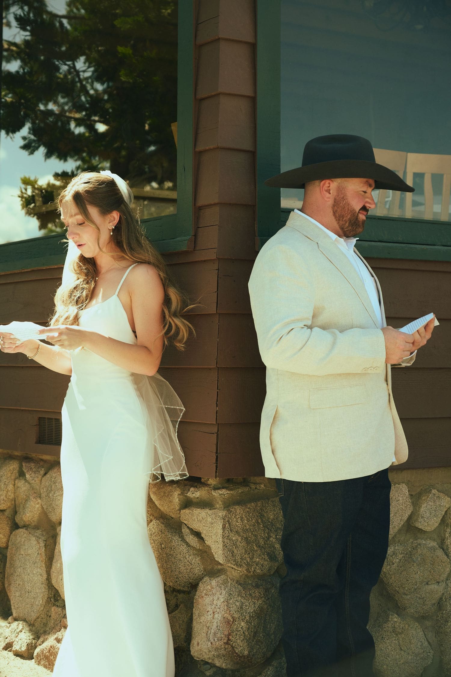 Bride in white gown and groom in cream suit with a black cowboy hat reading vows back-to-back at their Lake Tahoe wedding