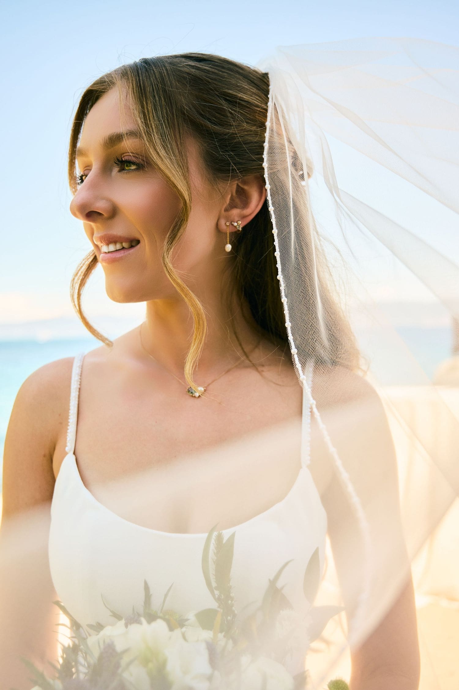 Smiling bride in white dress and veil holding a bouquet on the beach with a blue lake backdrop