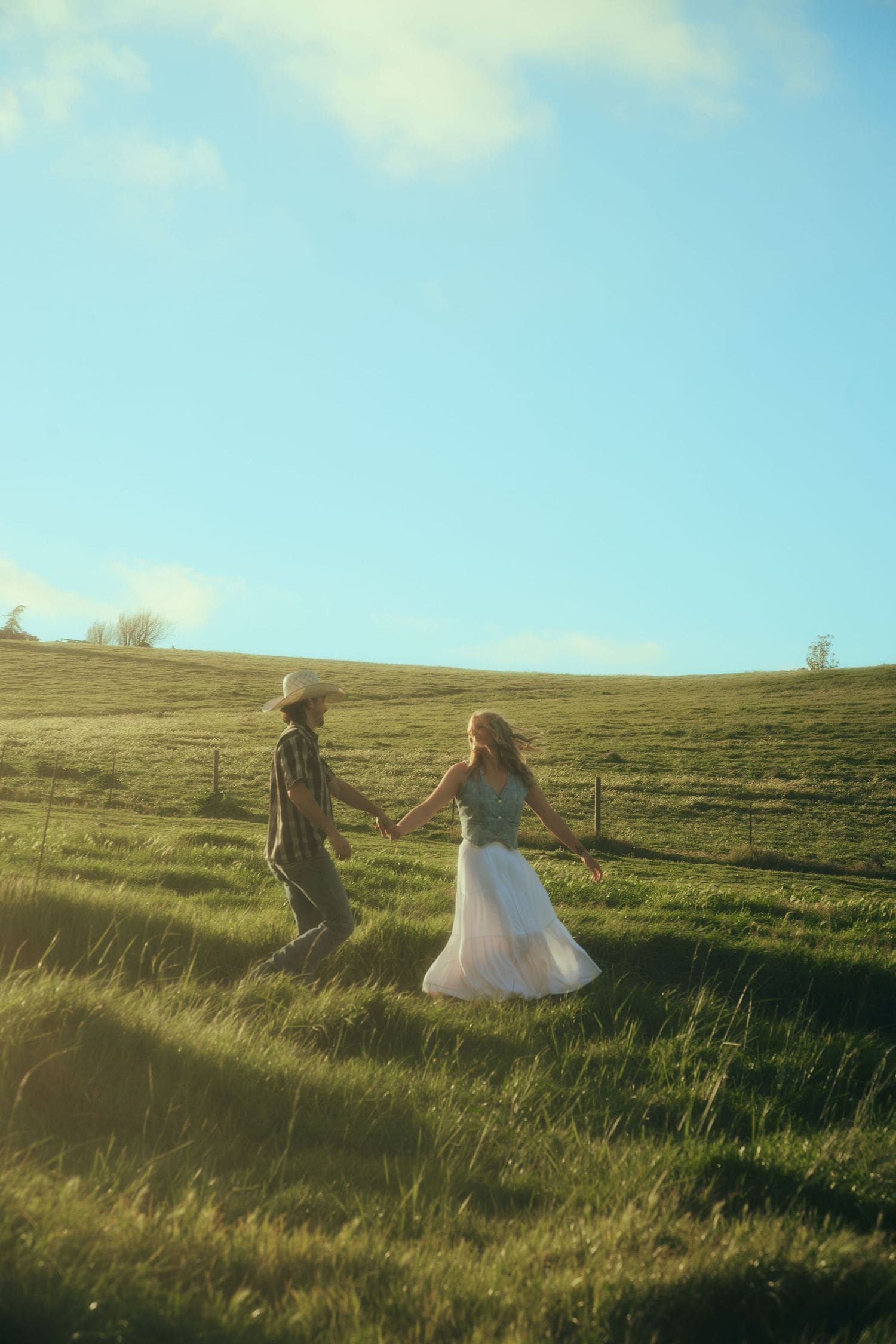 Couple holding hands walking through a sunny green meadow during a nostalgic countryside engagement session