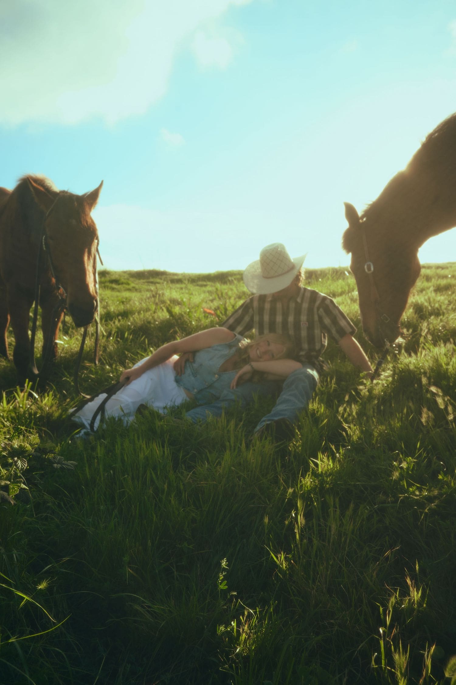 Engaged couple sitting in a lush green field surrounded by grazing horses under a bright blue sky