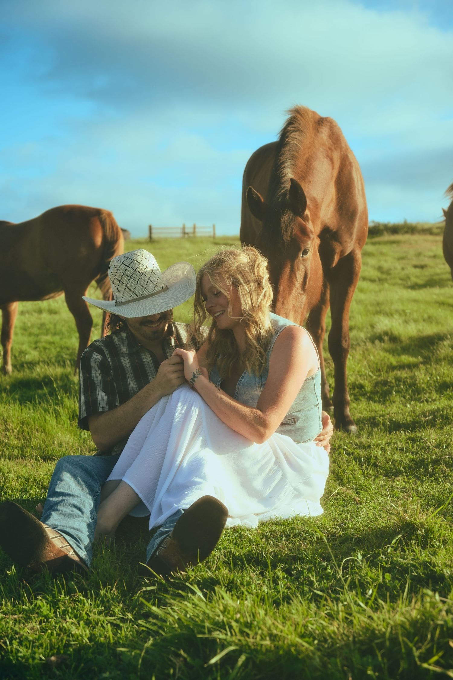 Couple sitting on green grass with brown horses grazing behind them during a countryside engagement session