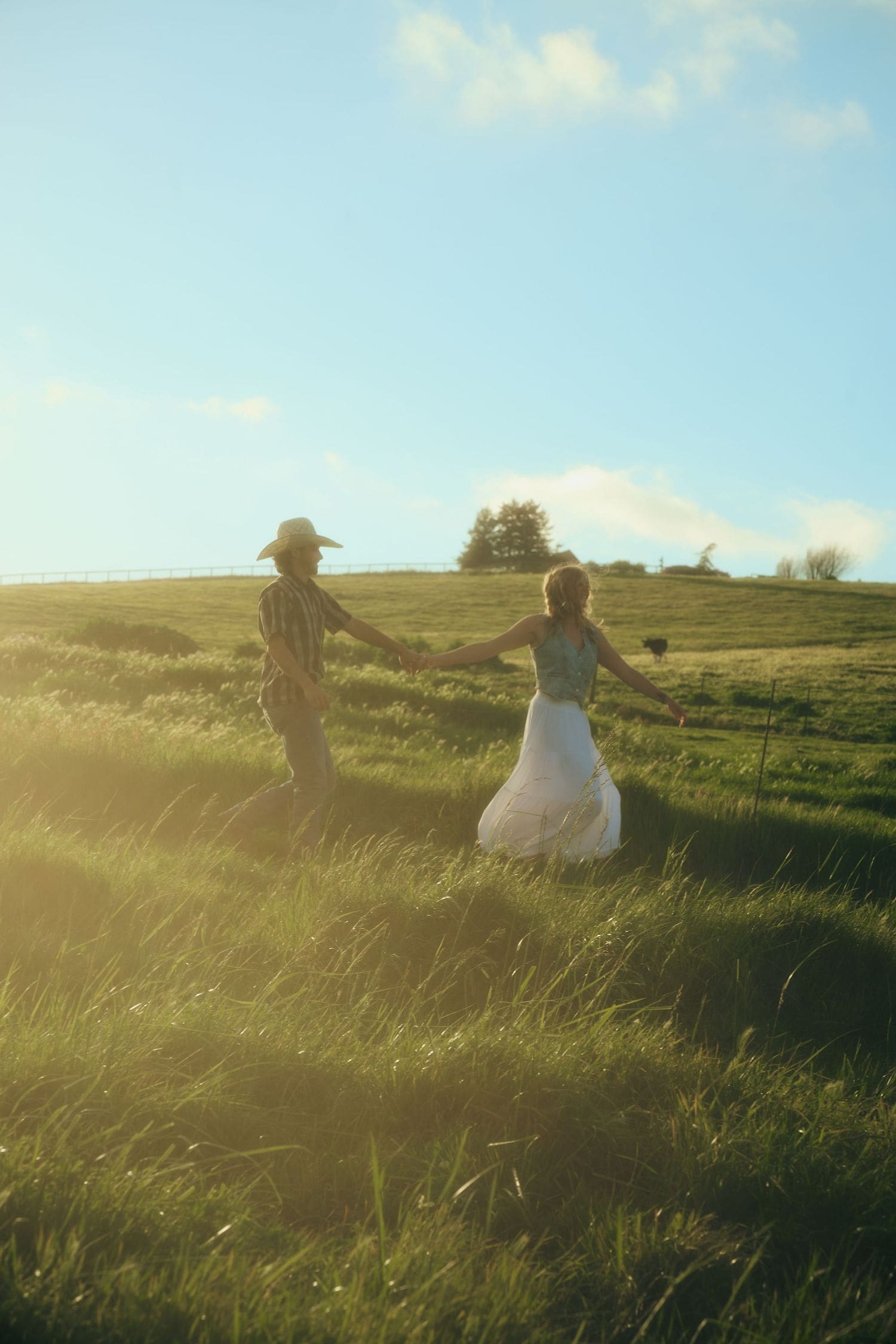Couple holding hands walking through a sunlit green meadow at their nostalgic engagement session