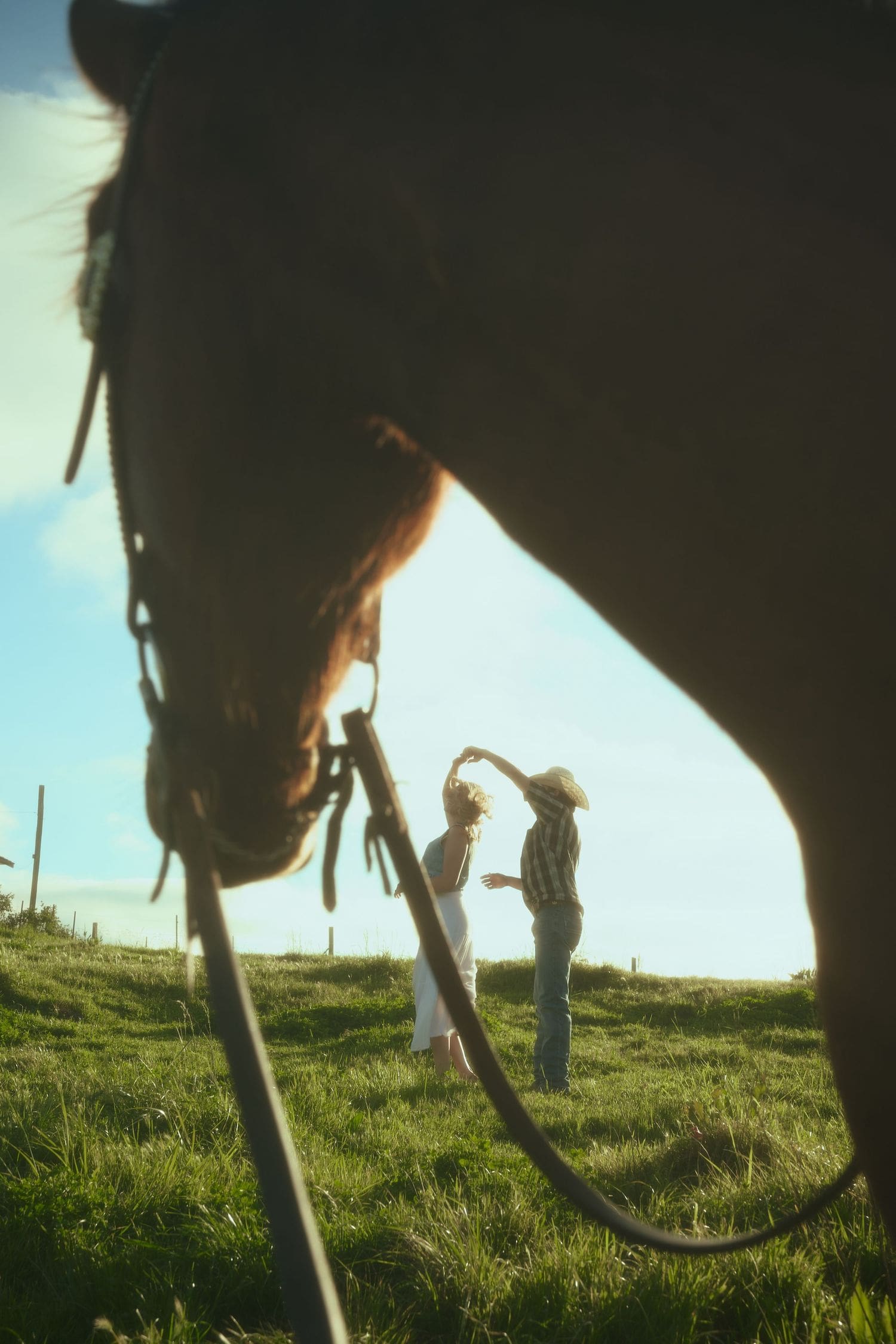 A horse frames a dancing couple on a sunny green hillside during a countryside engagement session