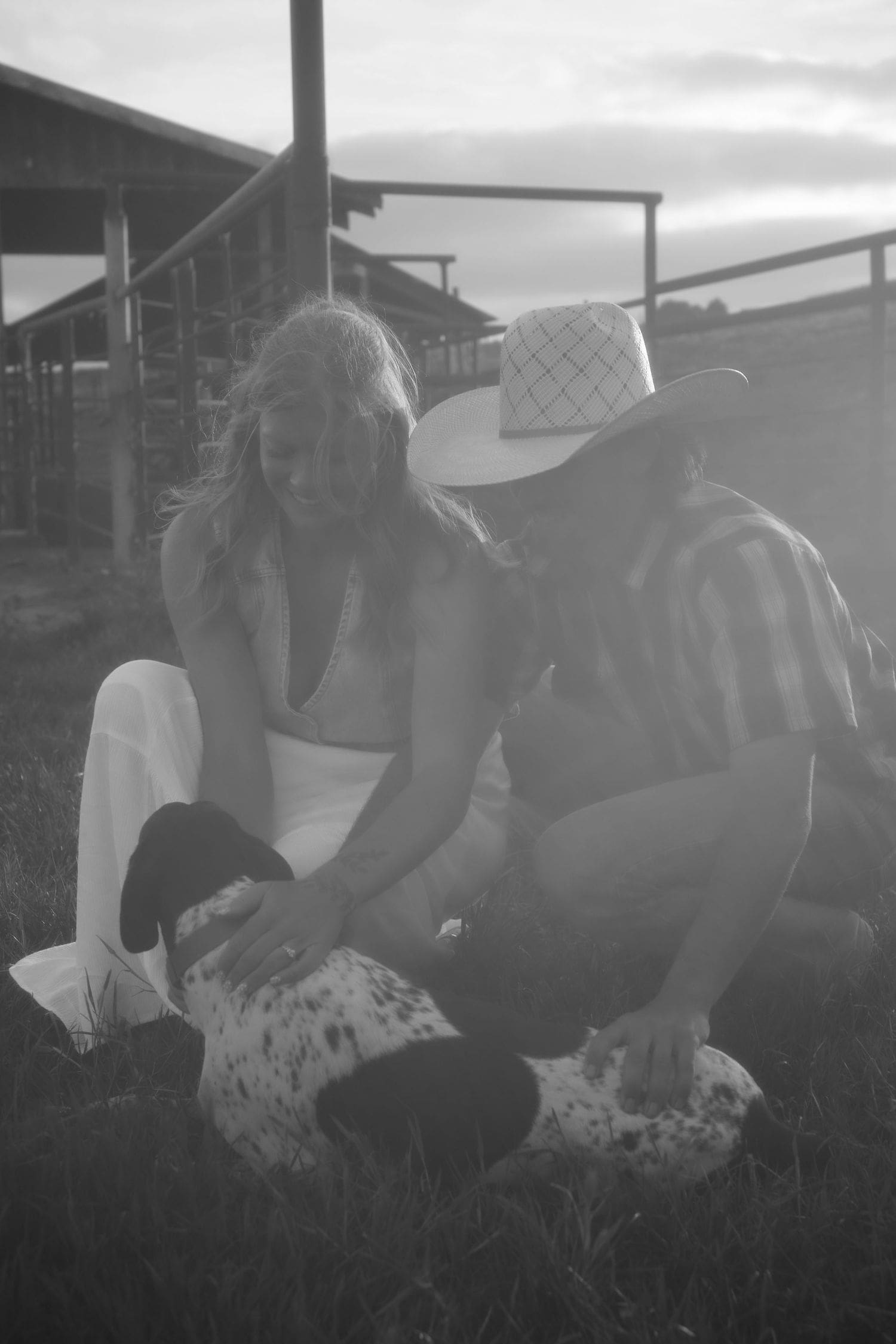 Black and white photo of a couple with a spotted dog near a farm fence during their engagement session